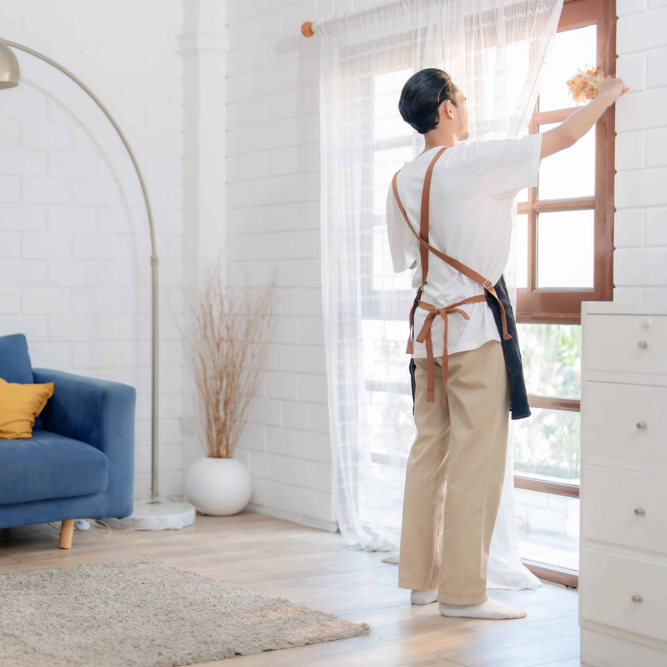 Homme en tablier faisant le ménage, époussetant le cadre d'une fenêtre en bois avec un plumeau marron dans un salon lumineux.
