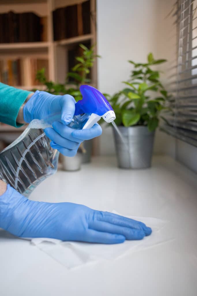 Désinfection surface avec gants : nettoyage efficace Personne portant des gants bleus désinfecte un comptoir blanc avec un vaporisateur bleu et essuie avec une lingette.