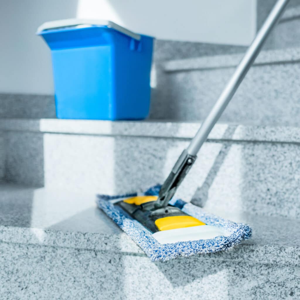 Nettoyage d'escalier en granit : serpillière et seau Homme nettoyant un escalier en granit avec une serpillière plate bleue et jaune, un seau bleu est visible en arrière-plan.