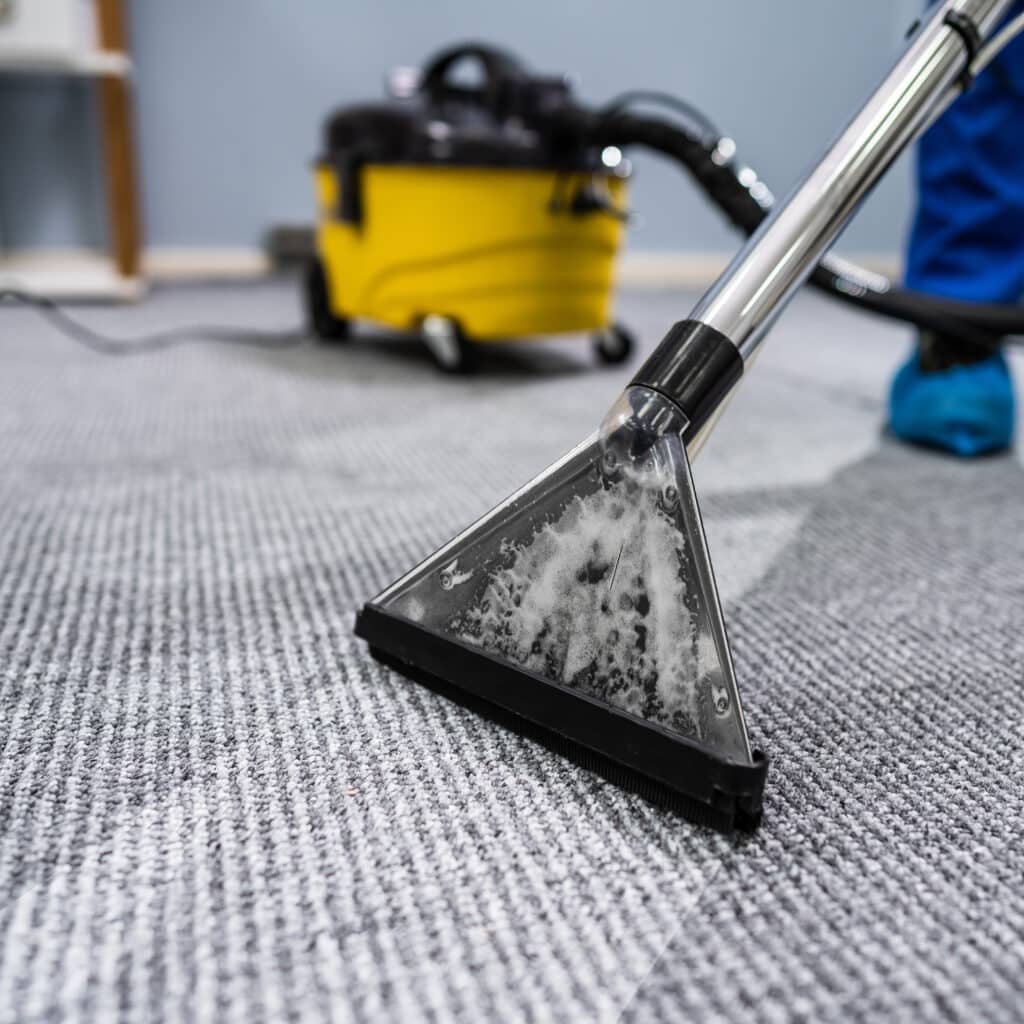 Nettoyage Professionnel de Moquette : Extraction à la Vapeur Nettoyage vapeur d'une moquette grise texturée avec une tête d'extraction professionnelle, mousse visible dans la buse.