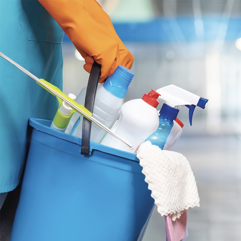 Produits de Nettoyage et Matériel pour Professionnels Professionnel portant un gant orange tenant un seau bleu rempli de produits de nettoyage, d'un raclette et d'un chiffon.